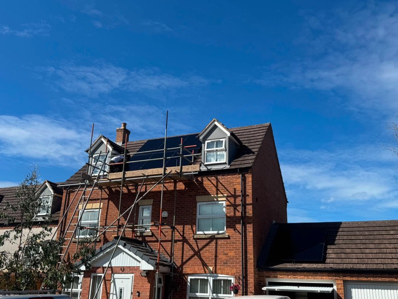 Solar panels being fitted to the roof of a three-storey brick house from scaffolding, under a clear blue sky